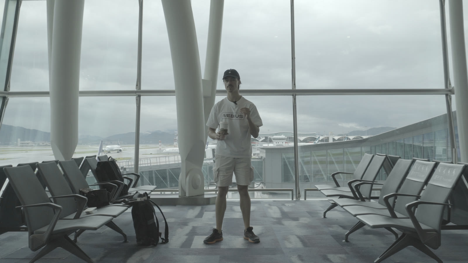 Ungraded S-Log2 footage of a man in a white T-shirt and cap at an overcast airport gate, backlit by tall windows: flat, grey, low contrast